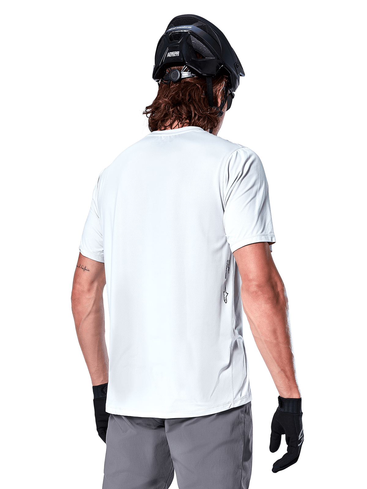 Alpinestars A-Dura Oscar Jersey, jersey de MTB de manga corta, White Dove, vista trasera de un ciclista con casco de ciclismo negro, pantalones cortos grises y guantes negros, equipamiento técnico de ciclismo de alto rendimiento