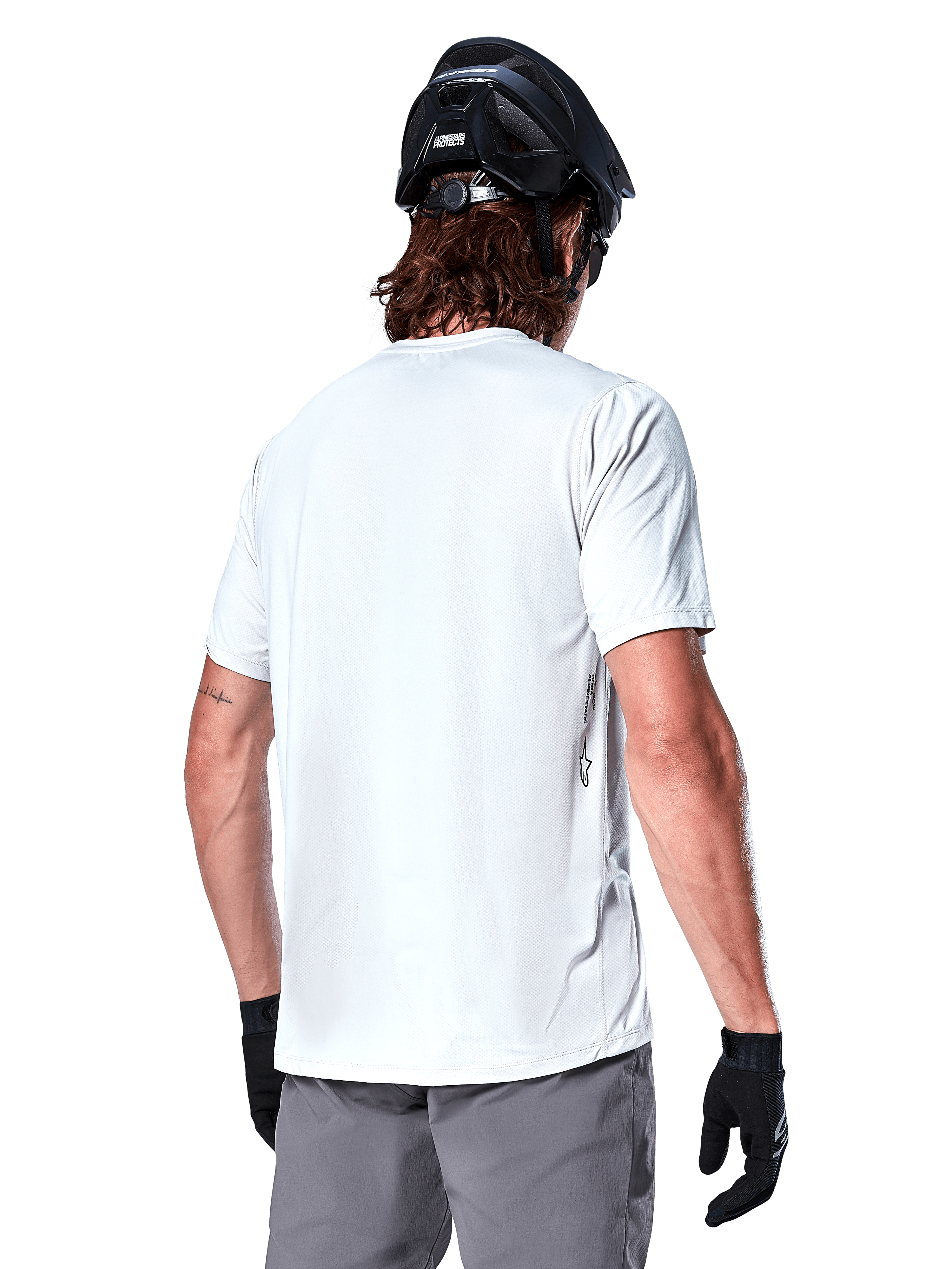 Alpinestars A-Dura Oscar Jersey, jersey de MTB de manga corta, White Dove, vista trasera de un ciclista con casco de ciclismo negro, pantalones cortos grises y guantes negros, equipamiento técnico de ciclismo de alto rendimiento