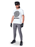 Alpinestars A-Dura Oscar Jersey - Manga Corta, White Dove con un gran gráfico de espiral negra, usado por un ciclista con pantalones de MTB grises, casco negro y guantes, ropa de ciclismo de alto rendimiento para rutas todoterreno