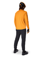 Ciclista de montaña visto desde atrás con el jersey de manga larga Alpinestars A-Dura Rocker en color Dark Gold, pantalones negros, casco naranja y negro, y zapatillas negras, equipamiento de MTB de alto rendimiento para recorrer senderos