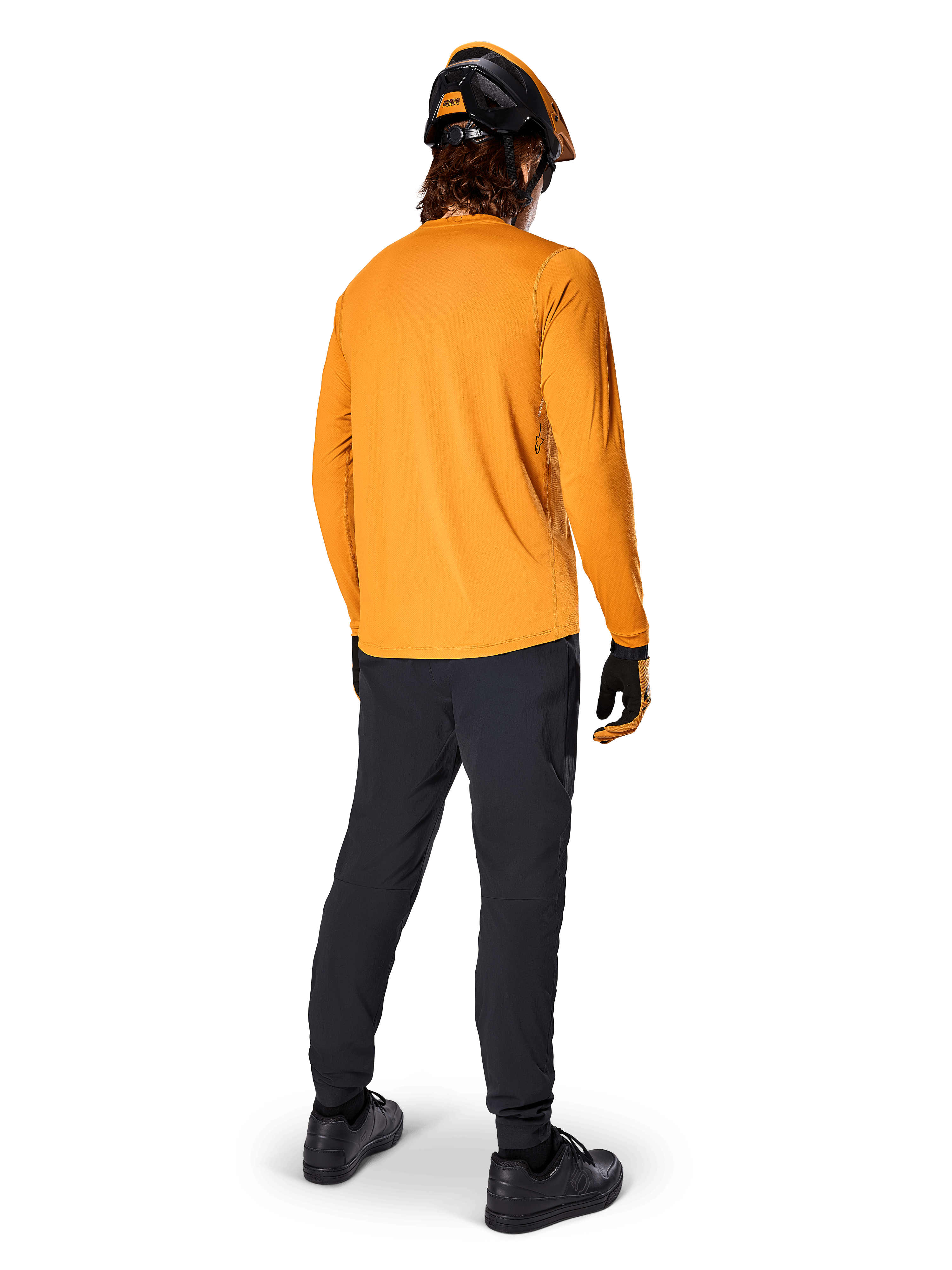 Ciclista de montaña visto desde atrás con el jersey de manga larga Alpinestars A-Dura Rocker en color Dark Gold, pantalones negros, casco naranja y negro, y zapatillas negras, equipamiento de MTB de alto rendimiento para recorrer senderos