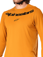 Camiseta de manga larga Alpinestars A-Dura Rocker, color Dark Gold, vista de primer plano del pecho con el logotipo y la marca Alpinestars en negro, tejido jacquard texturizado que absorbe la humedad, ropa técnica para ciclismo de montaña MTB