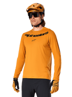 Alpinestars A-Dura Rocker Jersey de manga larga, camiseta para ciclismo de montaña MTB, color oro oscuro, persona con jersey, casco y guantes a juego, look deportivo coordinado con el logotipo de Alpinestars en el pecho