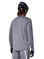 Jersey de manga larga Alpinestars A-Dura Rocker en gris oscuro, vista trasera de un ciclista de montaña, combinado con pantalones cortos grises a juego con logotipos en blanco, casco de MTB negro y guantes negros, equipamiento profesional de ciclismo todoterreno.