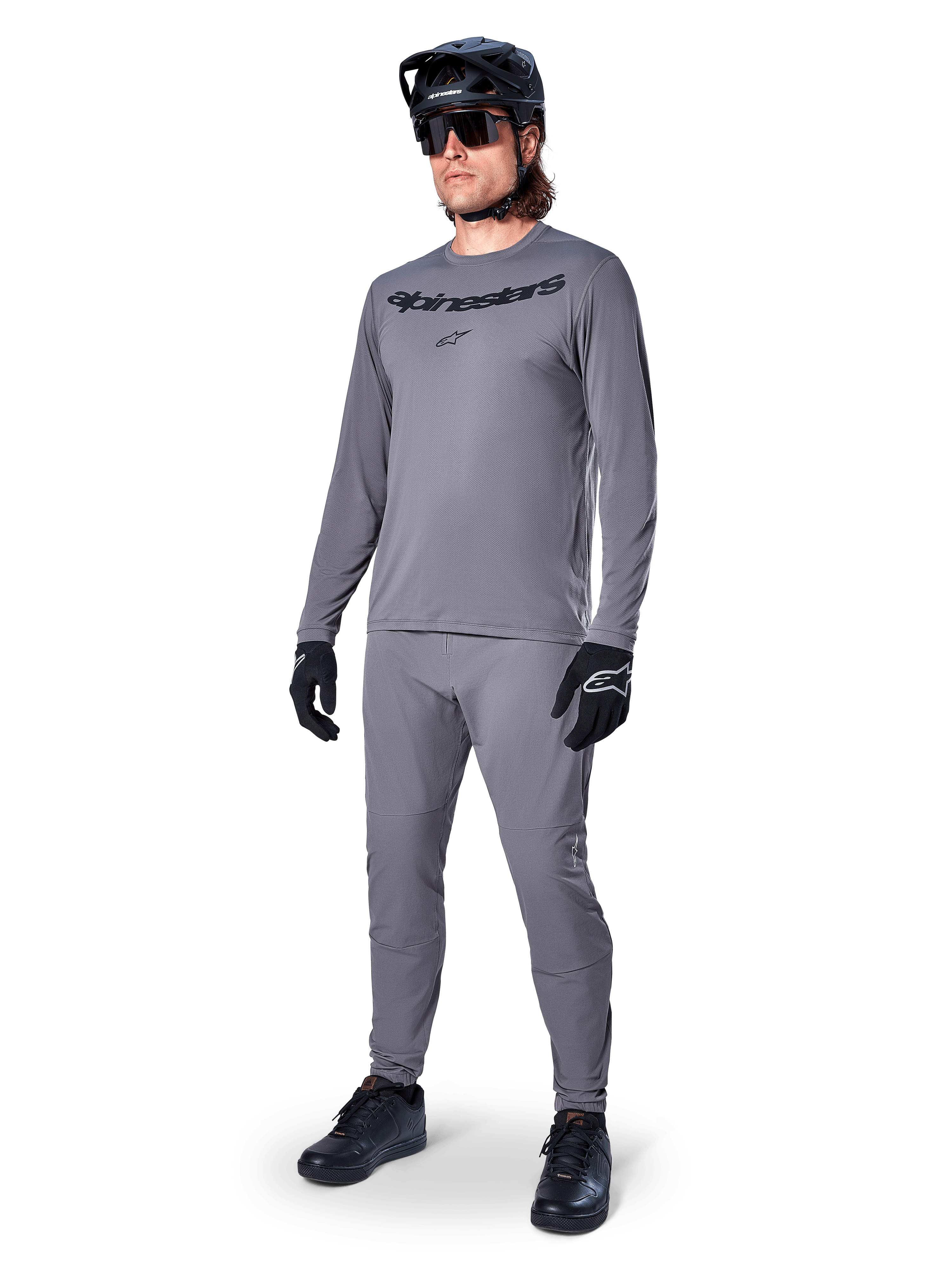 Alpinestars A-Dura Rocker Jersey - Long Sleeve, gris oscuro, usado por un modelo con pantalones grises a juego, casco de bicicleta de montaña negro, gafas de sol y guantes, estilo casual de MTB para conducción fuera de pista