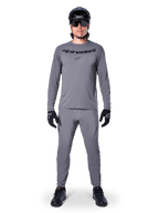 Ciclista masculino con el jersey de manga larga Alpinestars A-Dura Rocker en gris oscuro, pantalones grises a juego, casco de MTB negro, gafas de sol y guantes, equipamiento profesional de ciclismo de montaña para senderos todoterreno