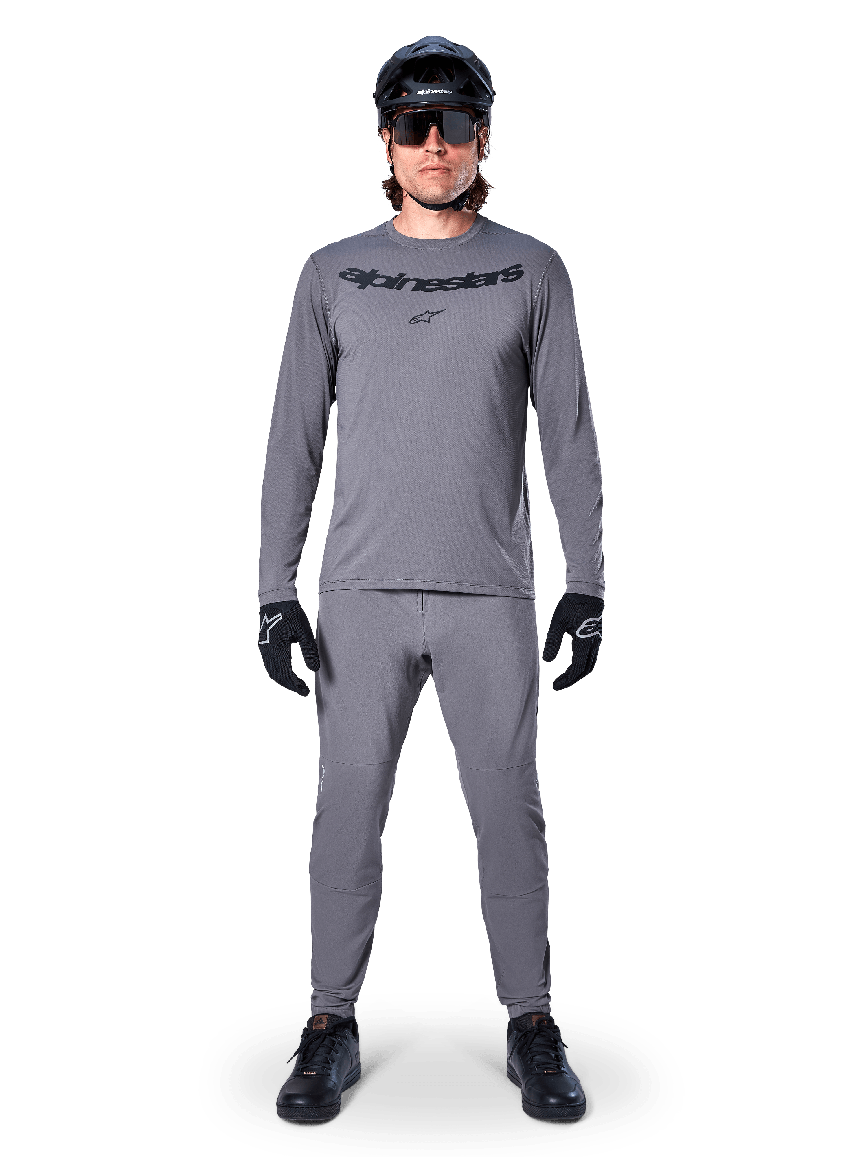 Ciclista masculino con el jersey de manga larga Alpinestars A-Dura Rocker en gris oscuro, pantalones grises a juego, casco de MTB negro, gafas de sol y guantes, equipamiento profesional de ciclismo de montaña para senderos todoterreno