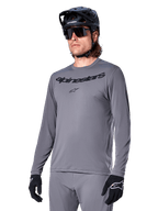 Alpinestars A-Dura Rocker Jersey de manga larga, jersey de MTB gris para ciclismo de montaña, usado con casco y guantes negros, con logotipo negro en el pecho, ropa deportiva de ciclismo para recorridos todoterreno, fondo liso