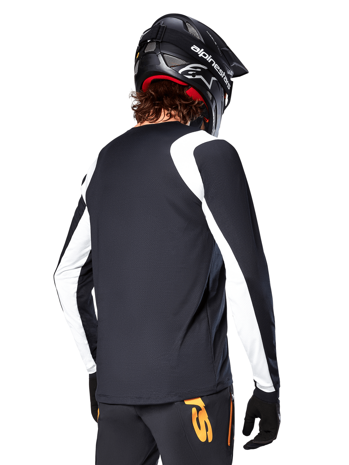 Ciclista de MTB con el Jersey de Manga Larga Alpinestars A-Supra Oscar, negro con paneles blancos en los hombros y las mangas, visto desde atrás, casco y guantes negros, pantalones con detalles naranjas, ropa de ciclismo todoterreno duradera.