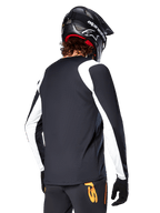 Ciclista de MTB con el Jersey de Manga Larga Alpinestars A-Supra Oscar, negro con paneles blancos en los hombros y las mangas, visto desde atrás, casco y guantes negros, pantalones con detalles naranjas, ropa de ciclismo todoterreno duradera.