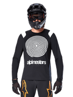 Jersey de manga larga Alpinestars A-Supra Oscar, negro con gráfico en espiral blanco y detalles en los hombros, usado por un ciclista con casco integral negro y gafas, pantalones negros y naranjas, equipamiento técnico de ciclismo para MTB de gravedad