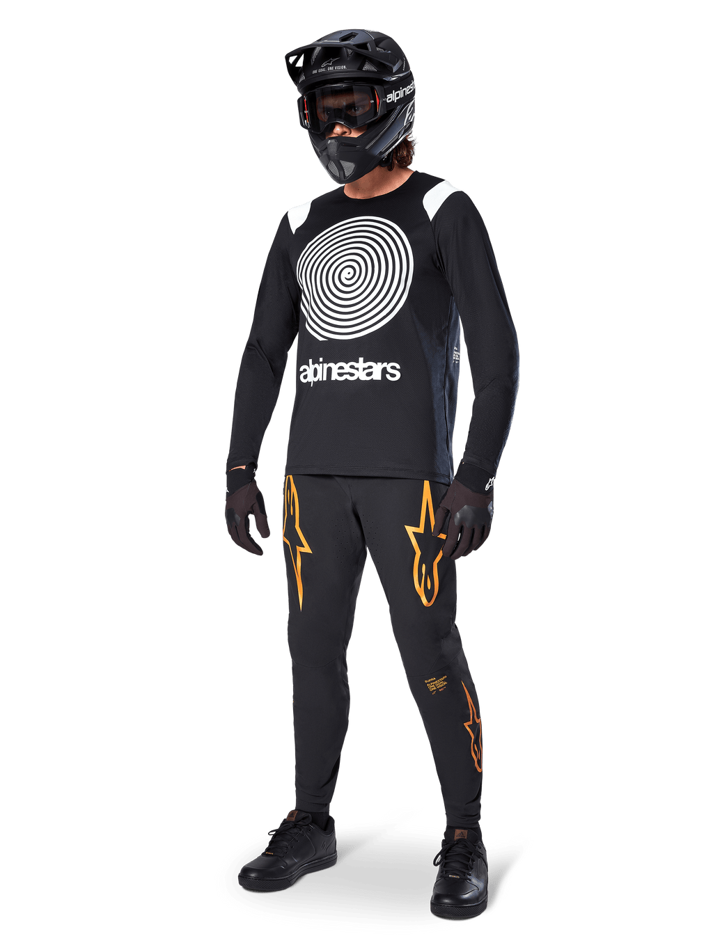 Ciclista de MTB con el jersey Alpinestars A-Supra Oscar de manga larga en color negro, con el gráfico de espiral Oscar vintage en blanco, pantalones negros con logotipos de estrellas naranjas, casco integral negro con gafas y guantes negros, equipamiento profesional para ciclismo de montaña