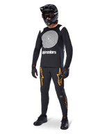 Ciclista de MTB con el jersey Alpinestars A-Supra Oscar de manga larga en color negro, con el gráfico de espiral Oscar vintage en blanco, pantalones negros con logotipos de estrellas naranjas, casco integral negro con gafas y guantes negros, equipamiento profesional para ciclismo de montaña