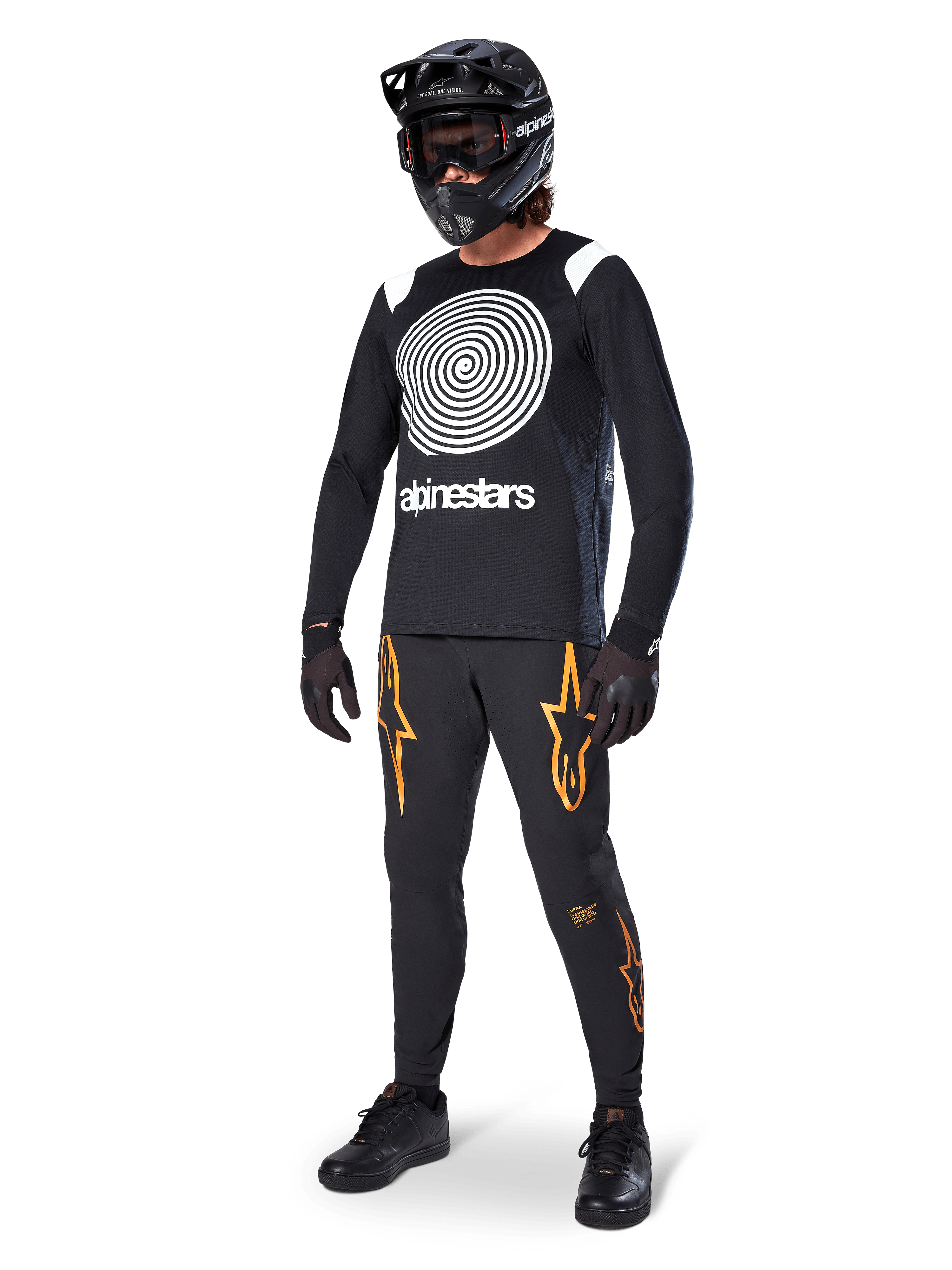 Ciclista de MTB con el jersey Alpinestars A-Supra Oscar de manga larga en color negro, con el gráfico de espiral Oscar vintage en blanco, pantalones negros con logotipos de estrellas naranjas, casco integral negro con gafas y guantes negros, equipamiento profesional para ciclismo de montaña
