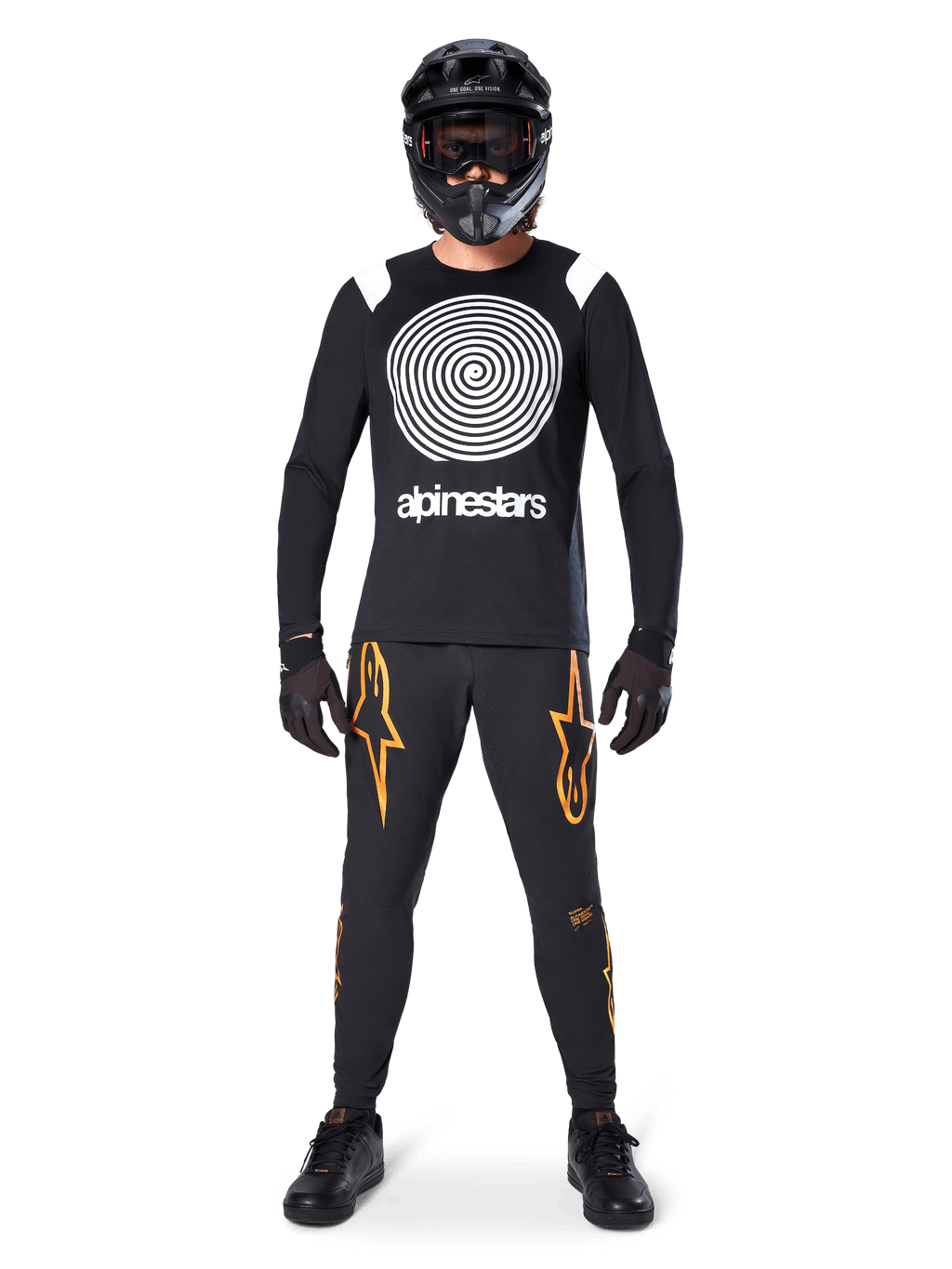 Modelo masculino con equipamiento completo para bicicleta de montaña, jersey de manga larga Alpinestars A-Supra Oscar en negro con gráfico de espiral blanco en el pecho, pantalones negros con logos de estrella naranjas, casco integral y gafas negras, guantes y zapatillas negras, atuendo coordinado para MTB de gravedad