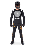 Modelo masculino con equipamiento completo para bicicleta de montaña, jersey de manga larga Alpinestars A-Supra Oscar en negro con gráfico de espiral blanco en el pecho, pantalones negros con logos de estrella naranjas, casco integral y gafas negras, guantes y zapatillas negras, atuendo coordinado para MTB de gravedad