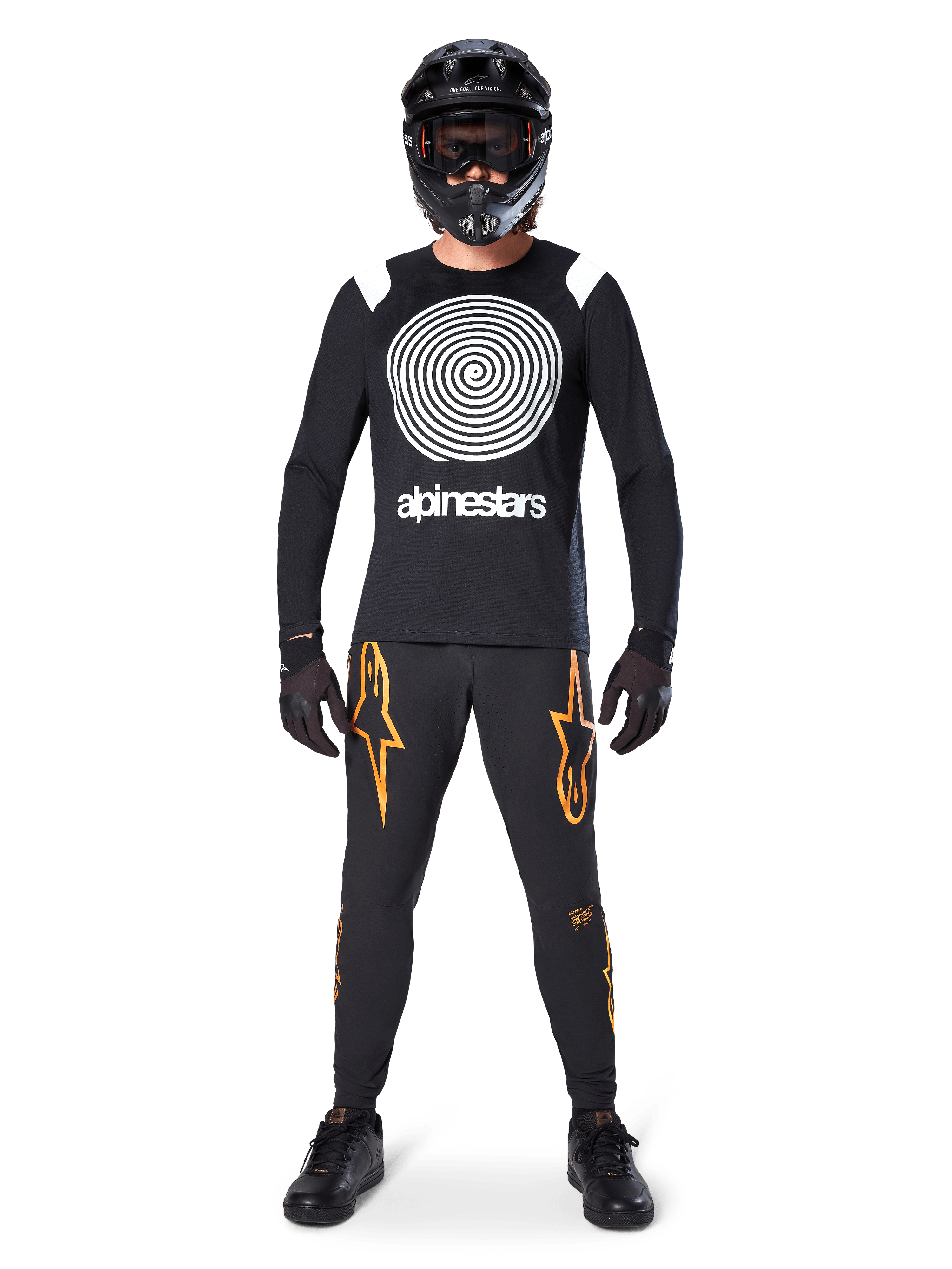Modelo masculino con equipamiento completo para bicicleta de montaña, jersey de manga larga Alpinestars A-Supra Oscar en negro con gráfico de espiral blanco en el pecho, pantalones negros con logos de estrella naranjas, casco integral y gafas negras, guantes y zapatillas negras, atuendo coordinado para MTB de gravedad