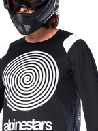 Jersey Alpinestars A-Supra Oscar de manga larga, negro, con un gran gráfico espiral Oscar blanco en el pecho y el logotipo de la marca, usado por un ciclista con casco integral negro y gafas, ropa técnica de mountain bike para gravity