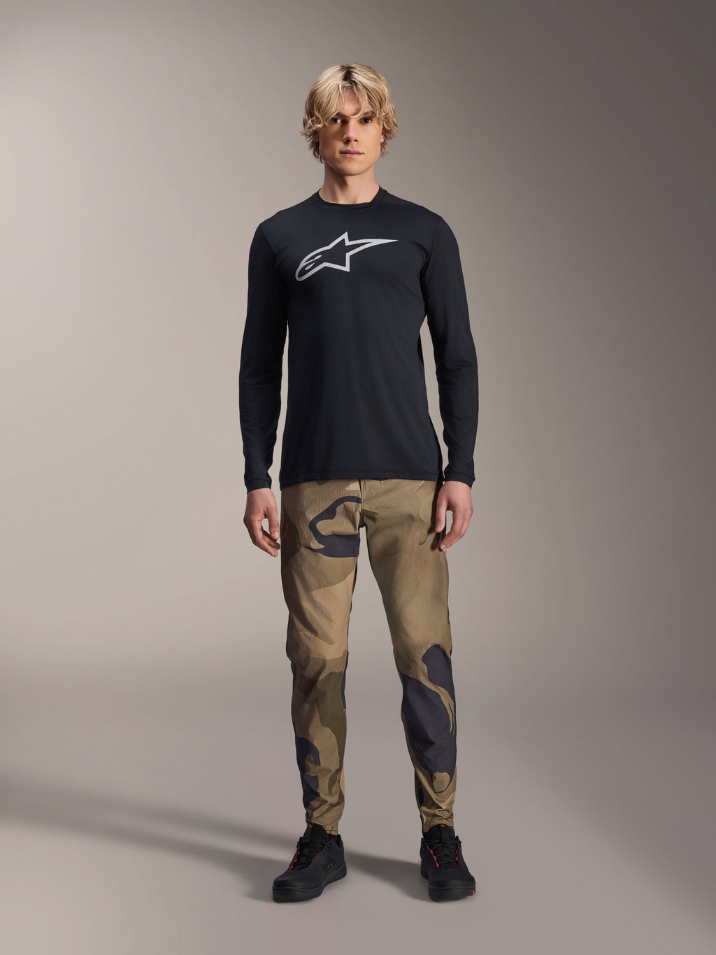 Alpinestars A-Dura Camo Pant, pantalones de ciclismo MTB, camuflaje marrón y verde, usados con un jersey negro de manga larga y zapatillas negras, estilo deportivo de montaña con tejido elástico duradero