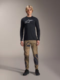 Alpinestars A-Dura Camo Pant, pantalones de ciclismo MTB, camuflaje marrón y verde, usados con un jersey negro de manga larga y zapatillas negras, estilo deportivo de montaña con tejido elástico duradero