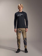 Alpinestars A-Dura Camo Pant, pantalones de ciclismo MTB, camuflaje marrón y verde, usados con un jersey negro de manga larga y zapatillas negras, estilo deportivo de montaña con tejido elástico duradero