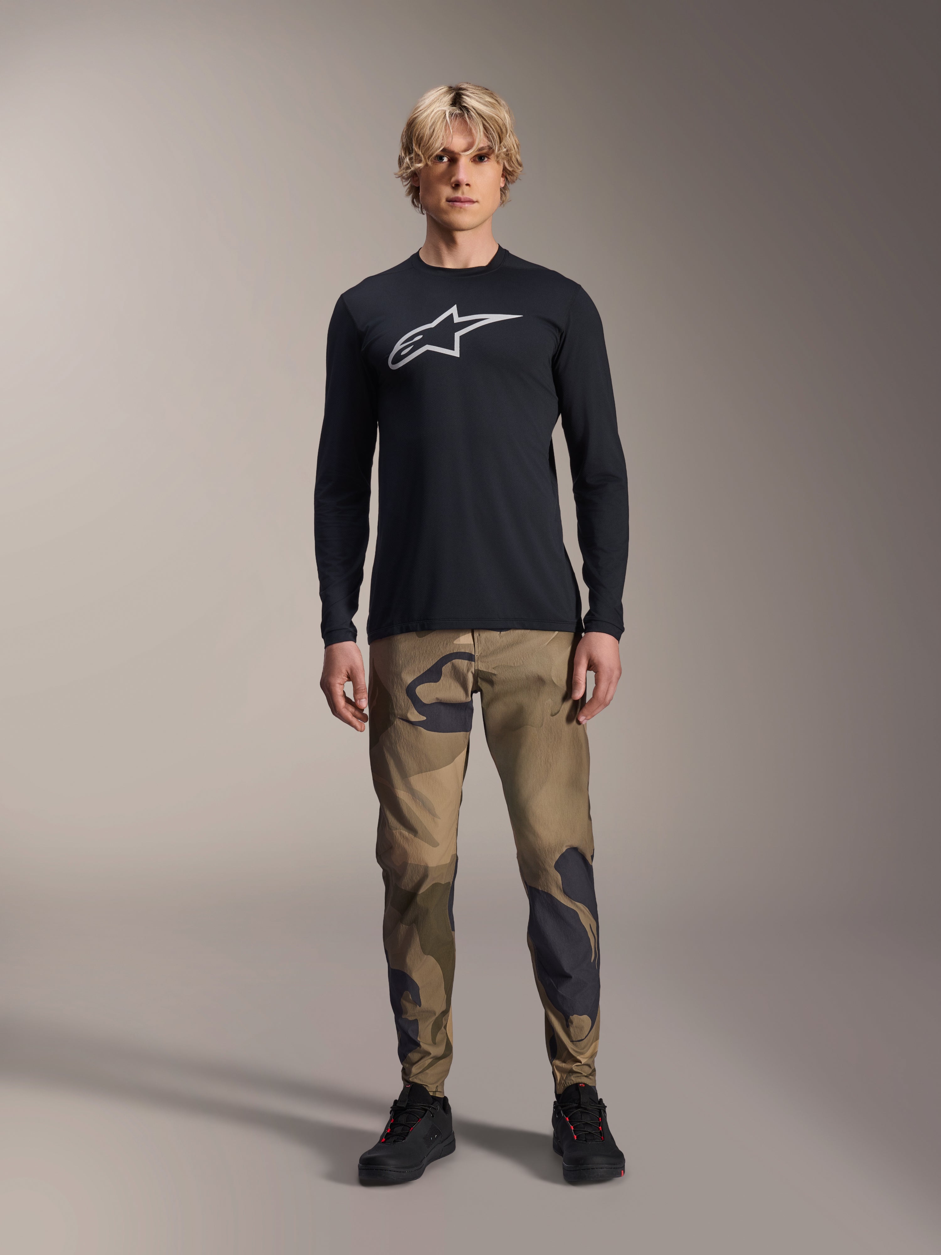 Alpinestars A-Dura Camo Pant, pantalones de ciclismo MTB, camuflaje marrón y verde, usados con un jersey negro de manga larga y zapatillas negras, estilo deportivo de montaña con tejido elástico duradero