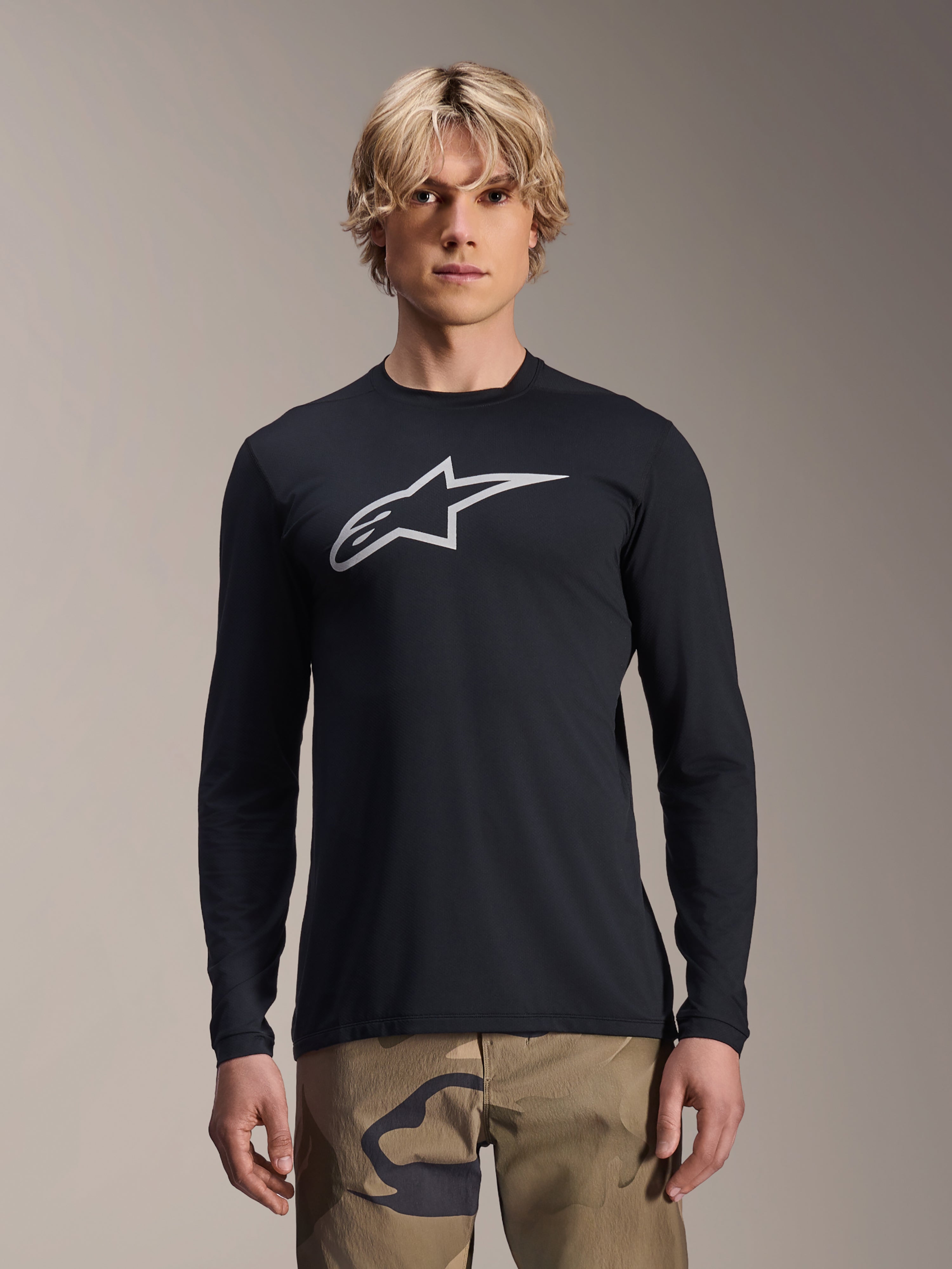 Jersey de manga larga Alpinestars A-Dura Astar, negro con logotipo plateado de Astars en el pecho, usado por un modelo con pantalones de camuflaje, ropa técnica casual para ciclismo de montaña MTB