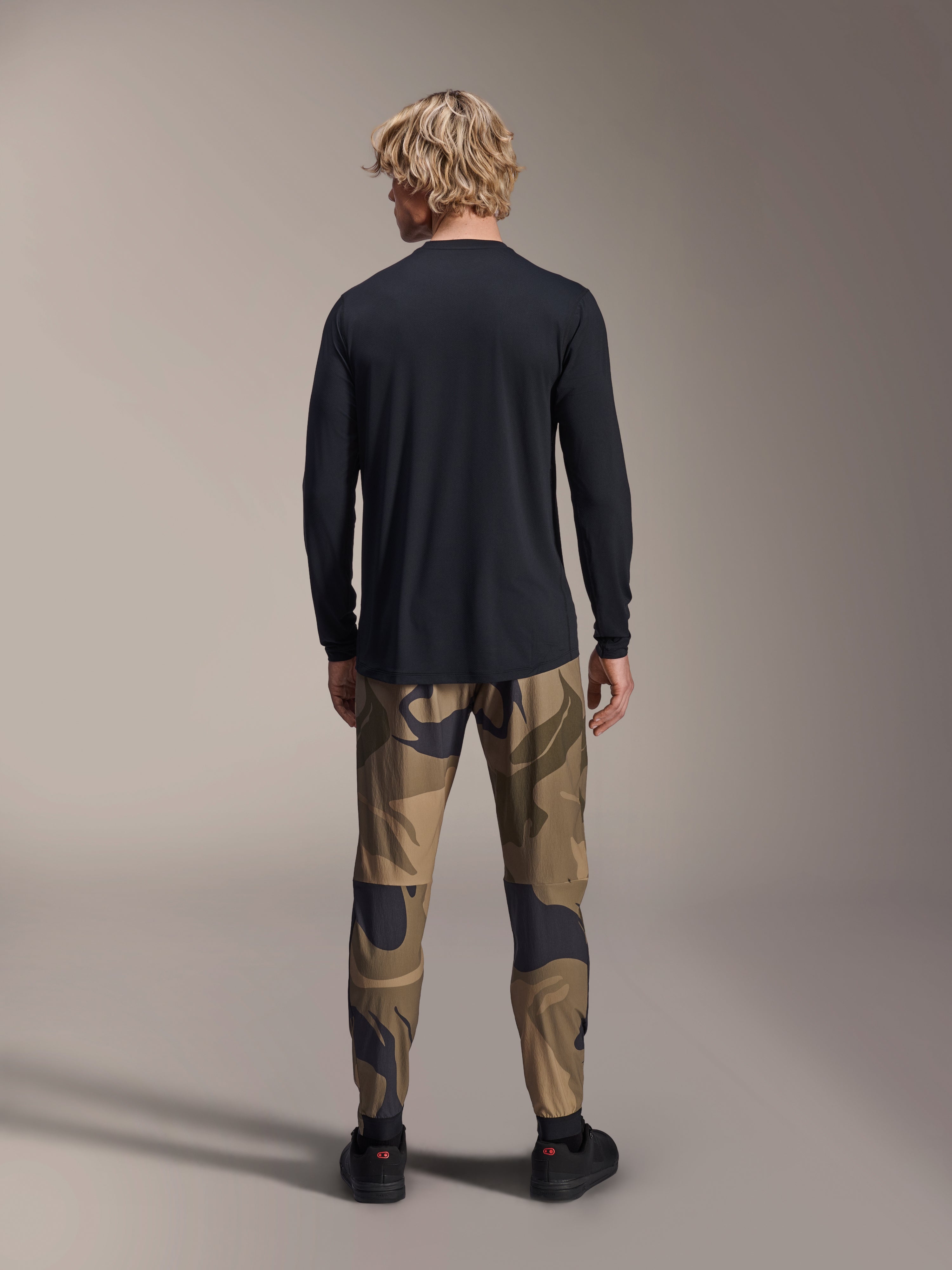 Pantalones Alpinestars A-Dura Camo, pantalones para bicicleta de montaña, patrón de camuflaje marrón, vista trasera, combinados con un jersey negro de manga larga y zapatillas negras, pantalones textiles de ajuste regular para ciclismo todoterreno