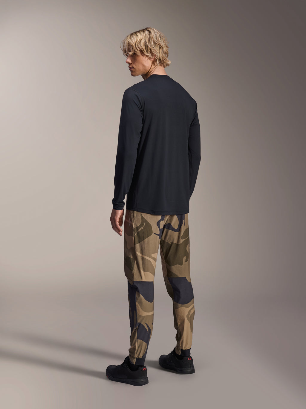 Alpinestars A-Dura Camo Pant, pantalones de MTB, camuflaje marrón, usados con un jersey negro de manga larga y zapatillas negras, vista trasera que muestra piernas ajustadas y puños elásticos, ropa técnica para ciclismo de montaña