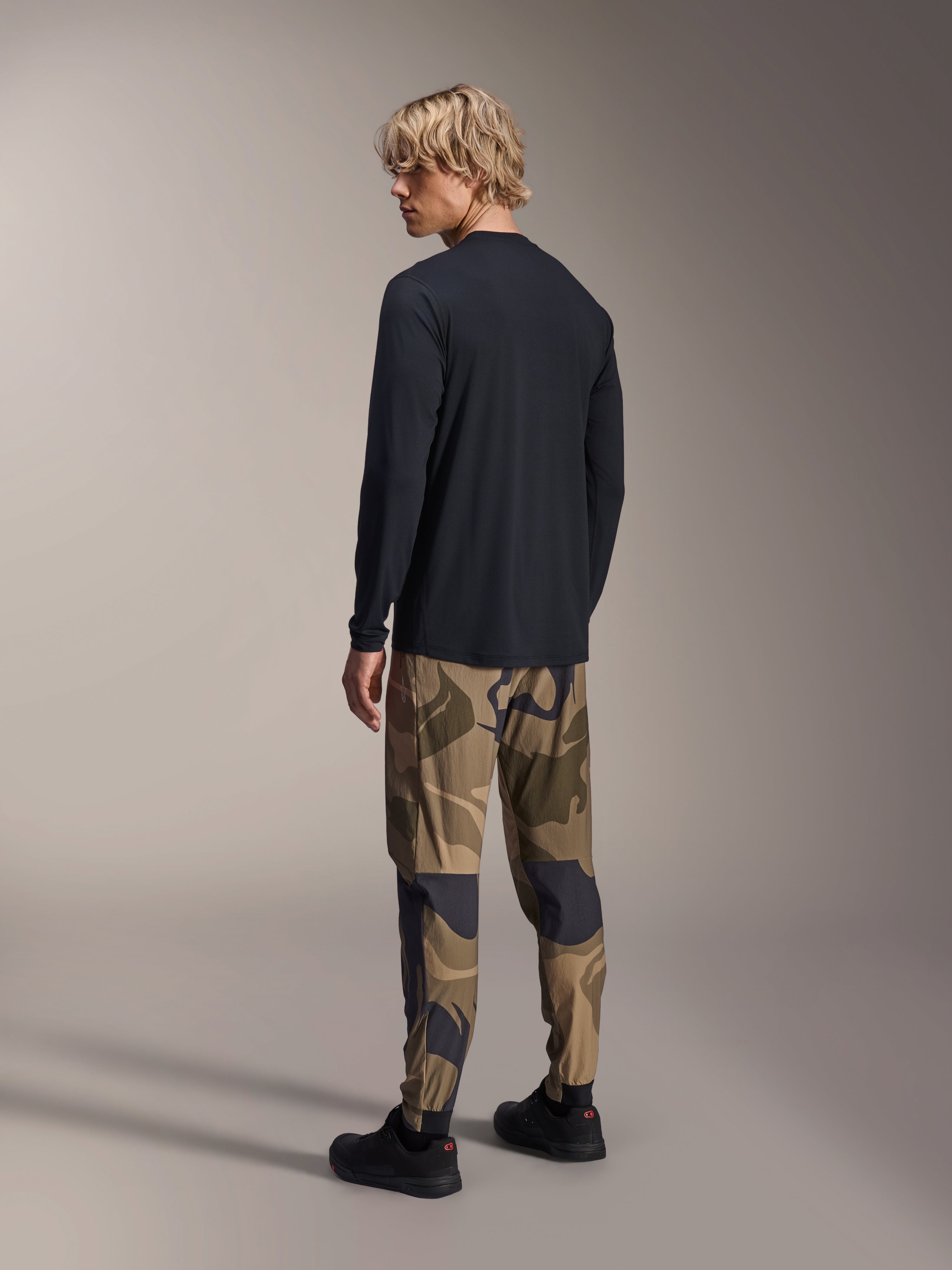 Alpinestars A-Dura Camo Pant, pantalones de MTB, camuflaje marrón, usados con un jersey negro de manga larga y zapatillas negras, vista trasera que muestra piernas ajustadas y puños elásticos, ropa técnica para ciclismo de montaña