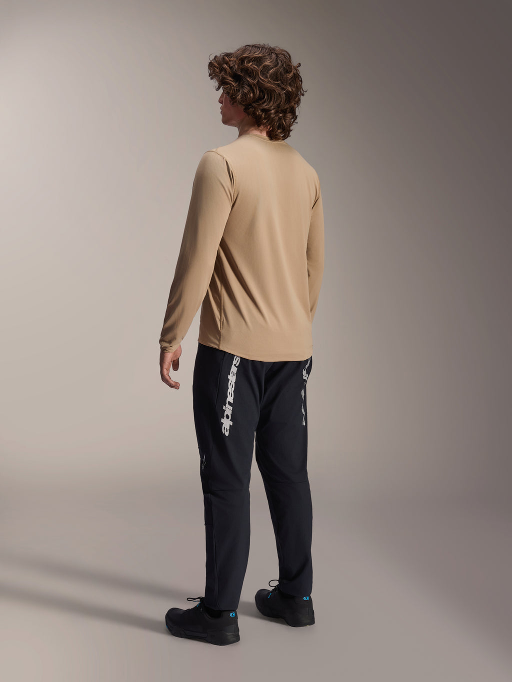 Alpinestars A-Dura Astar Long Sleeve Jersey in Light Brown, back view of a male model showcasing the drop tail design, paired with black pants and shoes, casual mountain bike performance gear.