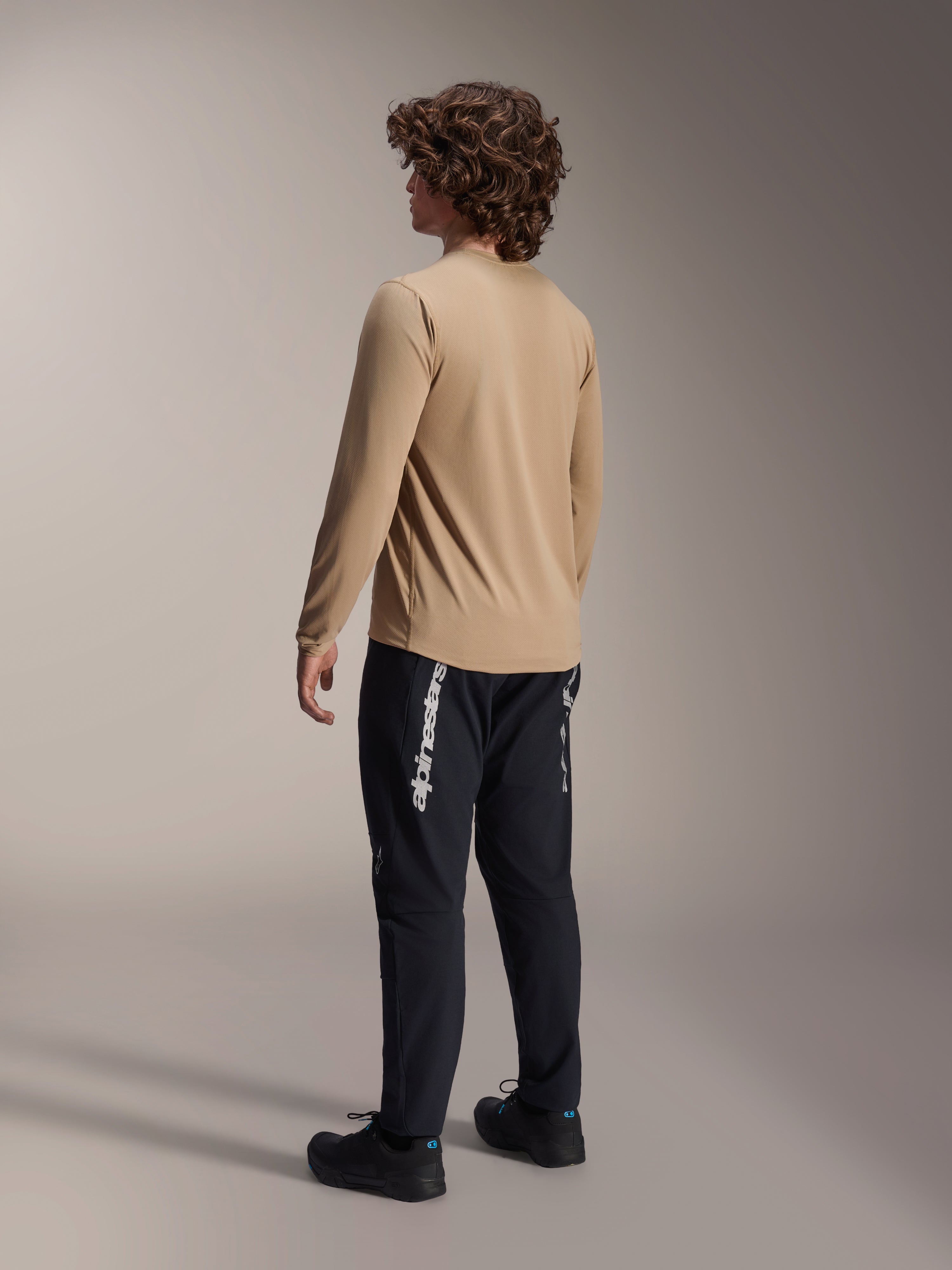 Alpinestars A-Dura Astar Long Sleeve Jersey in Light Brown, back view of a male model showcasing the drop tail design, paired with black pants and shoes, casual mountain bike performance gear.