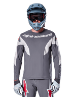 Ciclista de MTB vistiendo el Jersey Alpinestars A-Supra Race - Manga Larga en gris oscuro, jersey de ciclismo de manga larga con marca Alpinestars en blanco, combinado con un casco integral gris azulado, gafas y guantes de carreras grises, equipo completo de carreras de bicicleta de montaña para rendimiento todoterreno