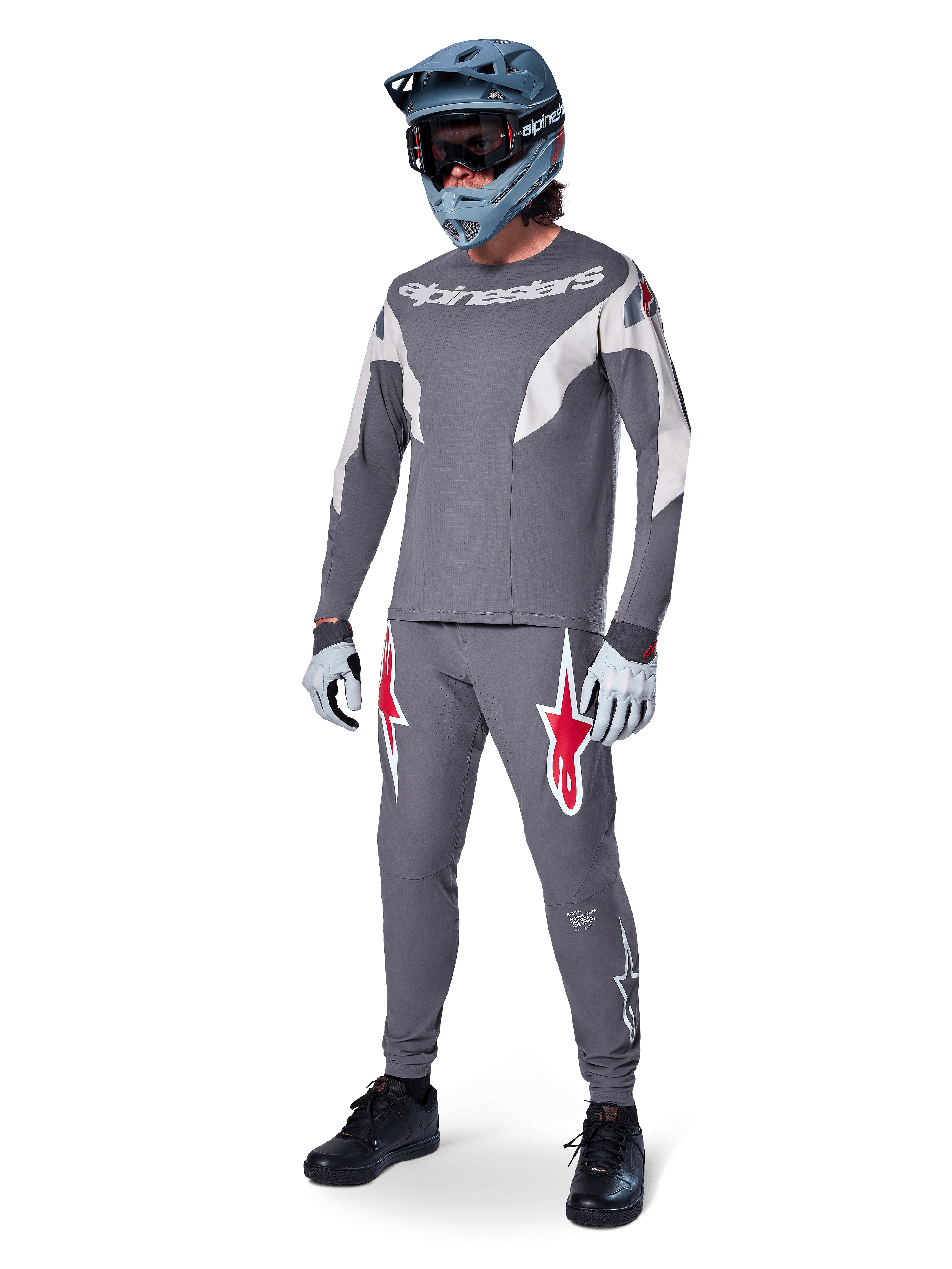 Ciclista de MTB con el Jersey Alpinestars A-Supra Race de manga larga, gris oscuro con detalles en gris claro, pantalones a juego con el logo de la estrella roja, casco integral gris con gafas y guantes gris claro, equipamiento coordinado para carreras de mountain bike
