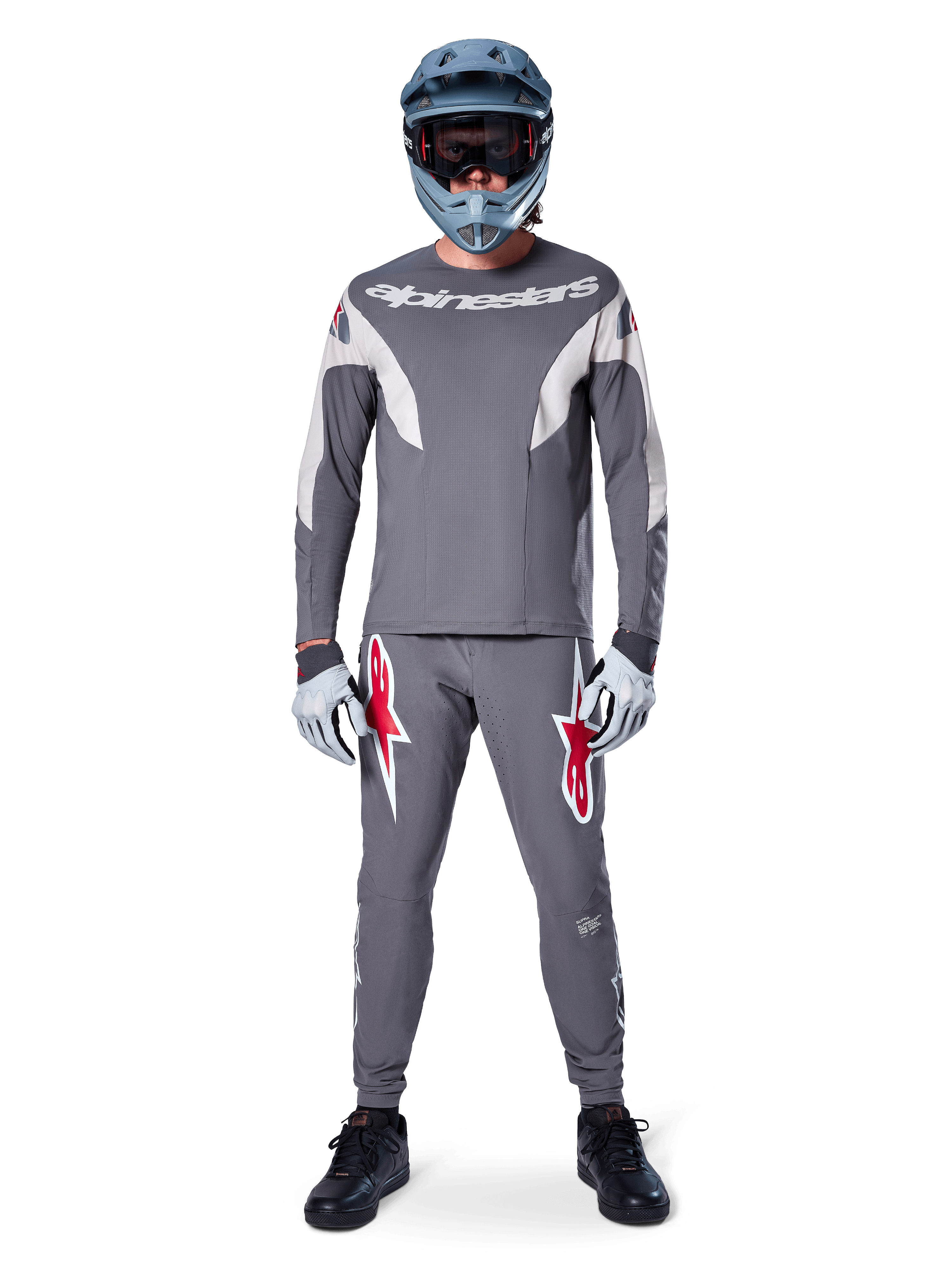 Piloto con el jersey de manga larga Alpinestars A-Supra Race en gris oscuro, combinado con pantalones grises a juego con llamativos logotipos de estrella rojos, casco gris con gafas y guantes gris claro, equipamiento completo de competición de MTB para ciclismo de montaña de alto rendimiento