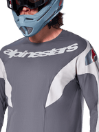 Alpinestars A-Supra Race Jersey - Manga Larga, jersey de MTB, gris oscuro con gráficos de carreras blancos y logotipo de Alpinestars blanco, usado con casco integral azul claro y gafas, tejido microperforado para rendimiento todoterreno