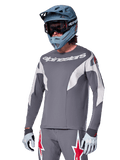 Alpinestars A-Supra Race Jersey de manga larga, gris oscuro con detalles blancos y rojos, gráficos de estilo de carreras, usado con pantalones grises a juego, casco integral azul y guantes blancos, equipo profesional de MTB y descenso
