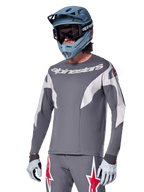 Alpinestars A-Supra Race Jersey de manga larga, gris oscuro con detalles blancos y rojos, gráficos de estilo de carreras, usado con pantalones grises a juego, casco integral azul y guantes blancos, equipo profesional de MTB y descenso
