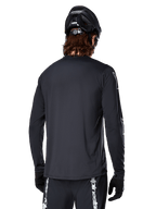Jersey de manga larga Alpinestars A-Dura Switch, negro, vista trasera de un ciclista con casco de MTB y guantes negros, con tejido jacquard técnico y parte trasera más larga para conducción off-road