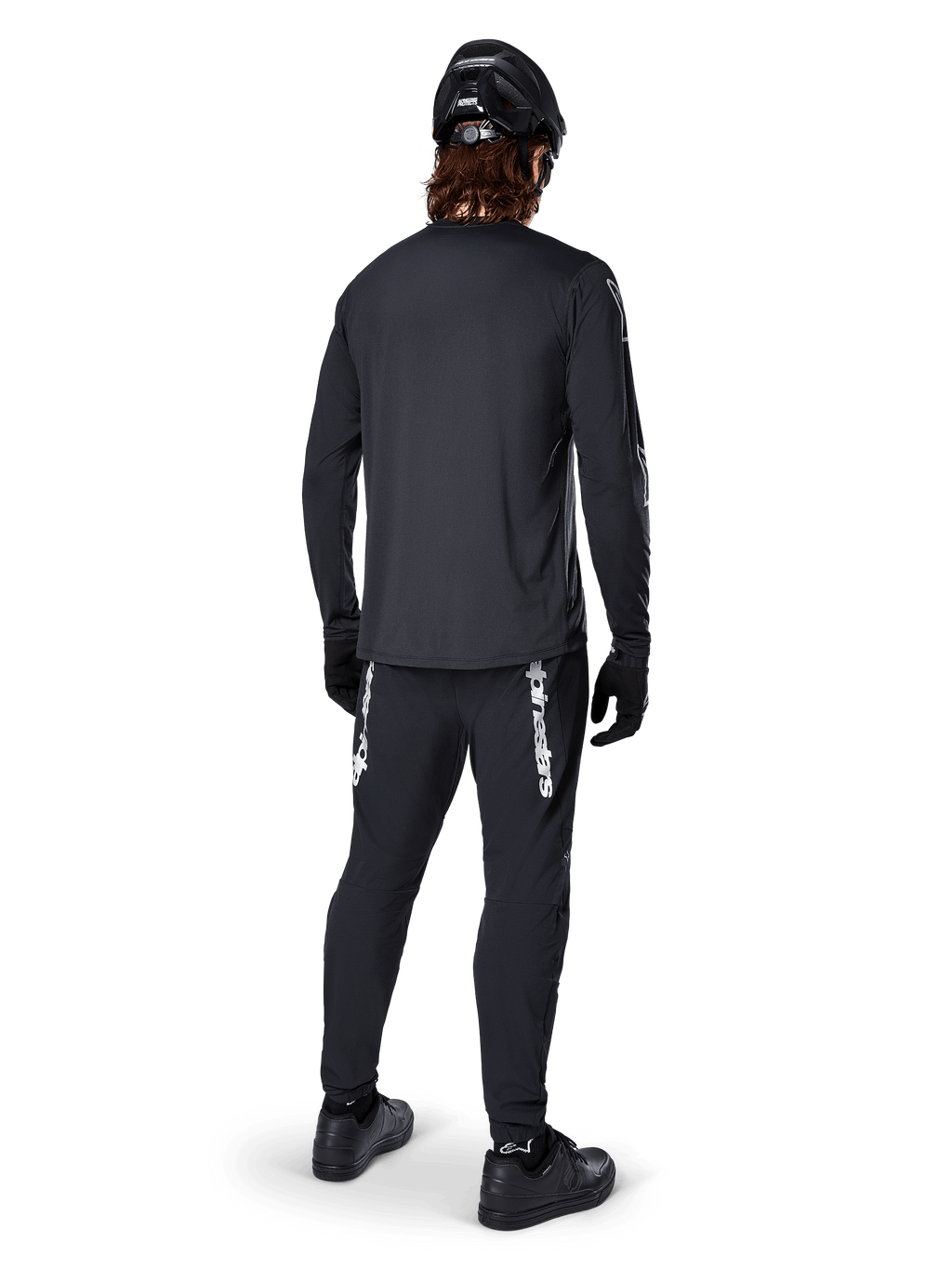 Jersey de manga larga Alpinestars A-Dura Switch, negro, vista trasera en un ciclista, usado con pantalones negros con la marca Alpinestars en blanco, casco y guantes negros, con una parte trasera más larga para mayor cobertura, ropa técnica para bicicleta de montaña