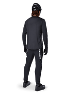 Jersey de manga larga Alpinestars A-Dura Switch, negro, vista trasera en un ciclista, usado con pantalones negros con la marca Alpinestars en blanco, casco y guantes negros, con una parte trasera más larga para mayor cobertura, ropa técnica para bicicleta de montaña