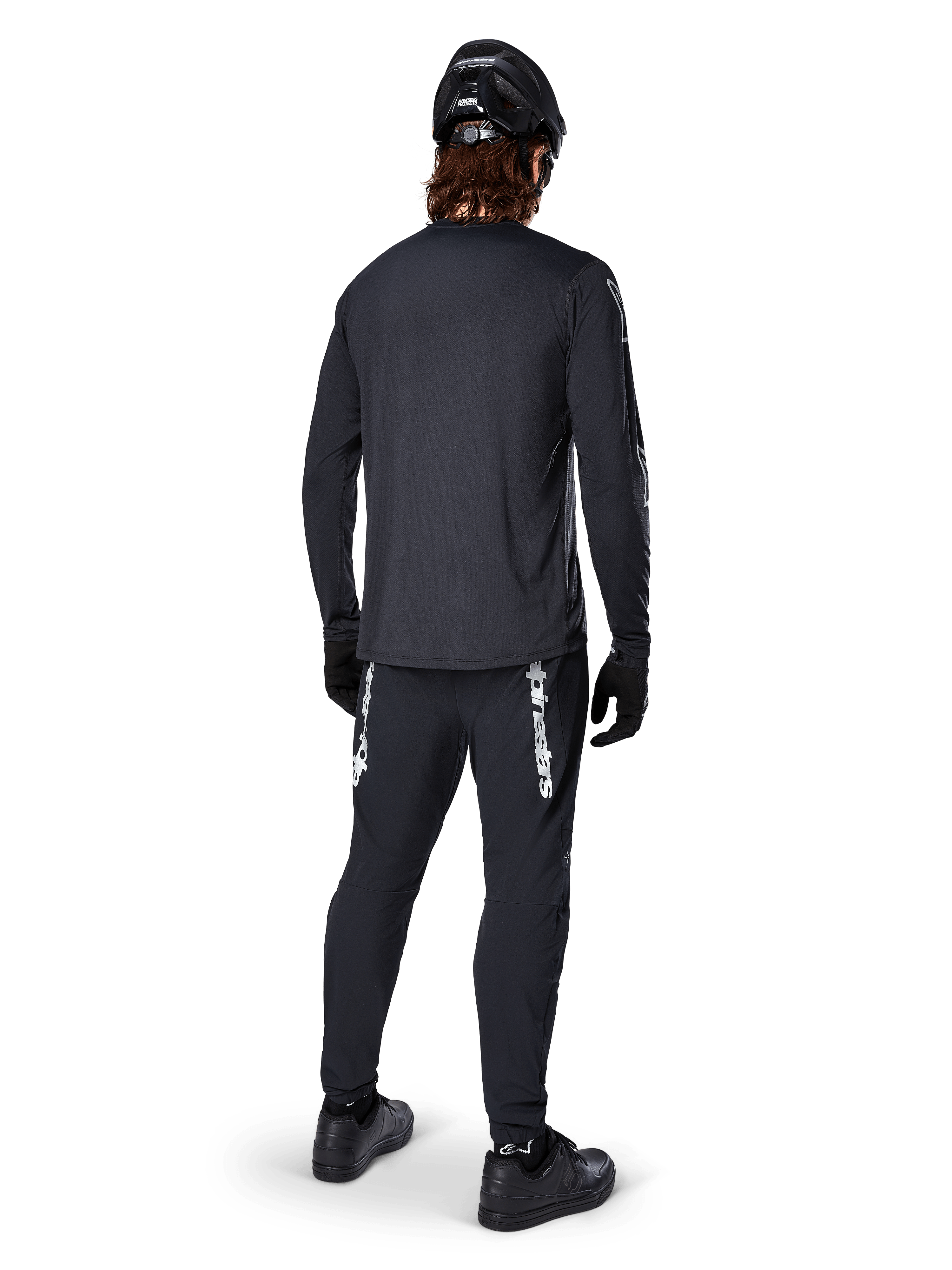 Jersey de manga larga Alpinestars A-Dura Switch, negro, vista trasera en un ciclista, usado con pantalones negros con la marca Alpinestars en blanco, casco y guantes negros, con una parte trasera más larga para mayor cobertura, ropa técnica para bicicleta de montaña