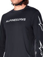 Jersey de manga larga Alpinestars A-Dura Switch, negro, con el logotipo de Alpinestars en blanco en el pecho y estrellas en la manga, usado por un ciclista con la correa del casco visible, ropa técnica para bicicleta de montaña