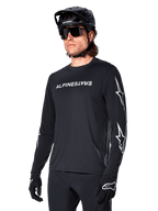Ciclista masculino con jersey de manga larga Alpinestars A-Dura Switch, negro con marca en blanco y logotipos de estrella en las mangas, combinado con casco de MTB negro y gafas de sol, equipamiento de ciclismo de alto rendimiento para rutas todoterreno