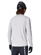 Jersey de manga larga Alpinestars A-Dura Switch, Gris Claro, vista trasera de un ciclista de montaña con casco de MTB negro y guantes, con una parte trasera más larga para mayor cobertura y tejido jacquard texturizado, equipamiento de ciclismo de alto rendimiento.