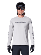 Alpinestars A-Dura Switch Long Sleeve Jersey, gris claro, presenta el nombre de la marca en gris oscuro en el pecho y logotipos de estrellas en las mangas, usado por un ciclista con casco de MTB y guantes negros, ropa de ciclismo de alto rendimiento para trail