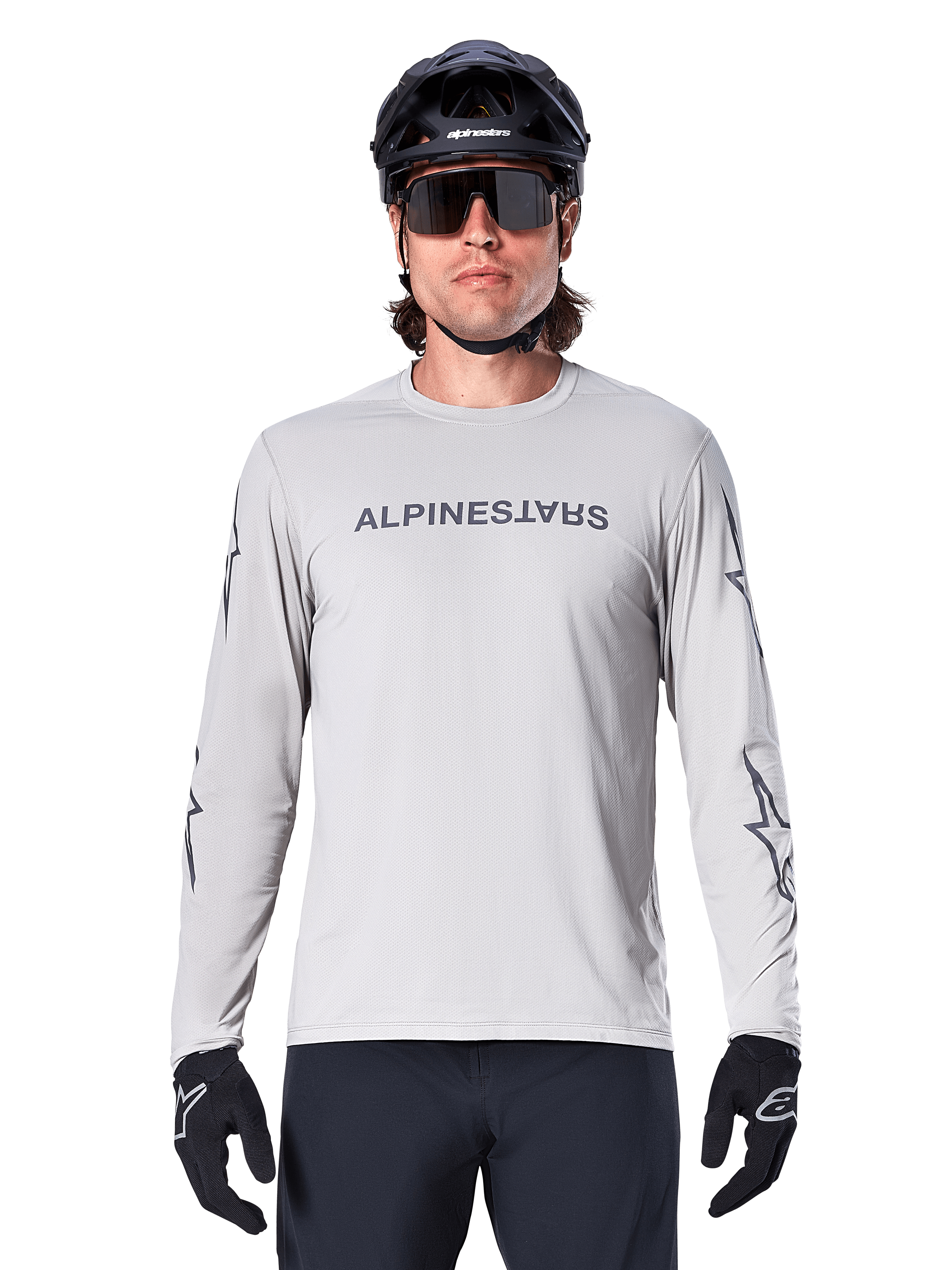 Alpinestars A-Dura Switch Long Sleeve Jersey, gris claro, presenta el nombre de la marca en gris oscuro en el pecho y logotipos de estrellas en las mangas, usado por un ciclista con casco de MTB y guantes negros, ropa de ciclismo de alto rendimiento para trail