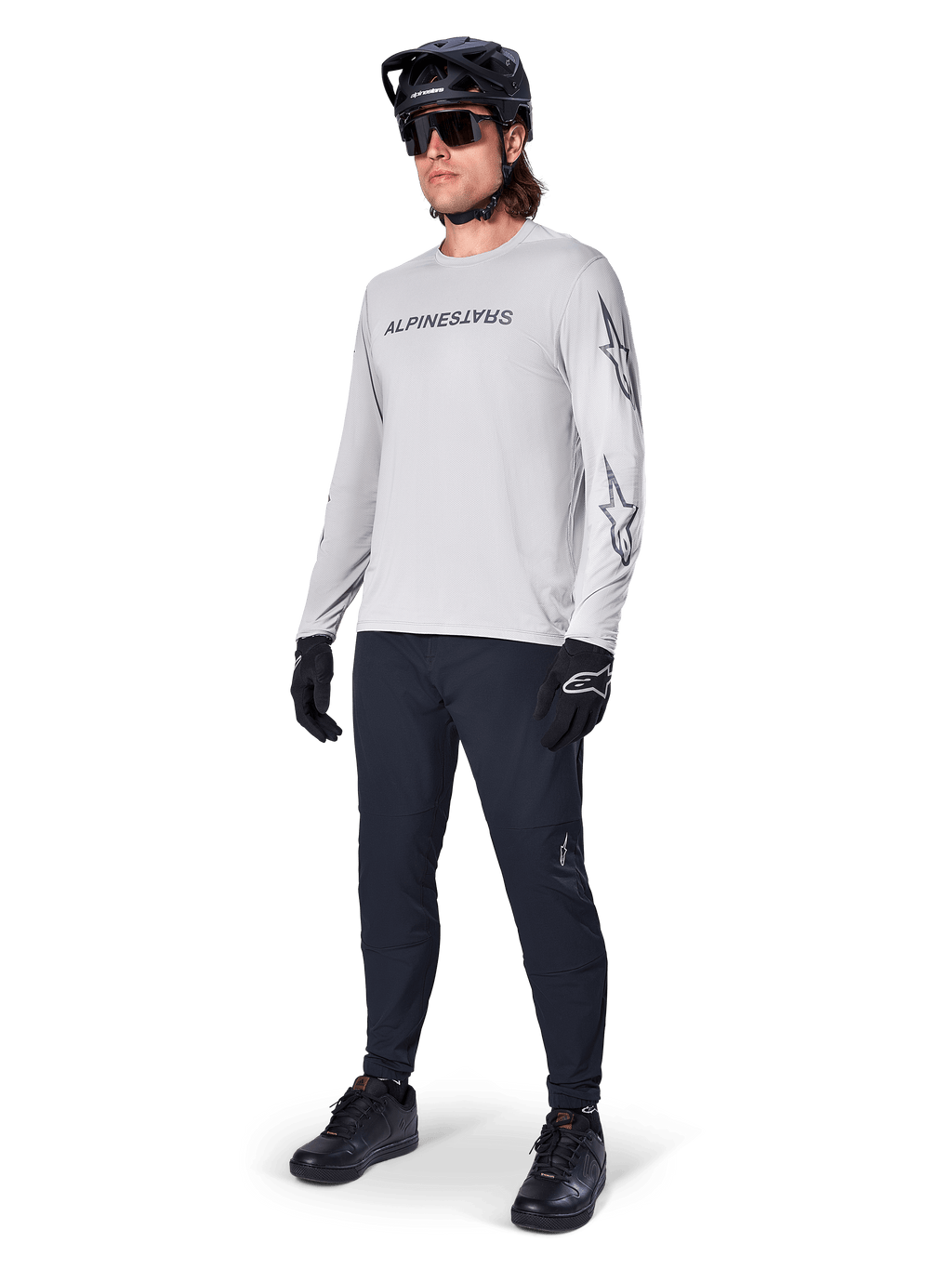 Jersey de manga larga Alpinestars A-Dura Switch, gris claro, usado por un ciclista con pantalones negros de montaña y casco, con logotipo de la marca en el pecho y gráficos de estrellas en las mangas, equipamiento de alto rendimiento para ciclismo todoterreno