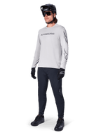 Jersey de manga larga Alpinestars A-Dura Switch, gris claro, usado por un ciclista con pantalones negros de montaña y casco, con logotipo de la marca en el pecho y gráficos de estrellas en las mangas, equipamiento de alto rendimiento para ciclismo todoterreno
