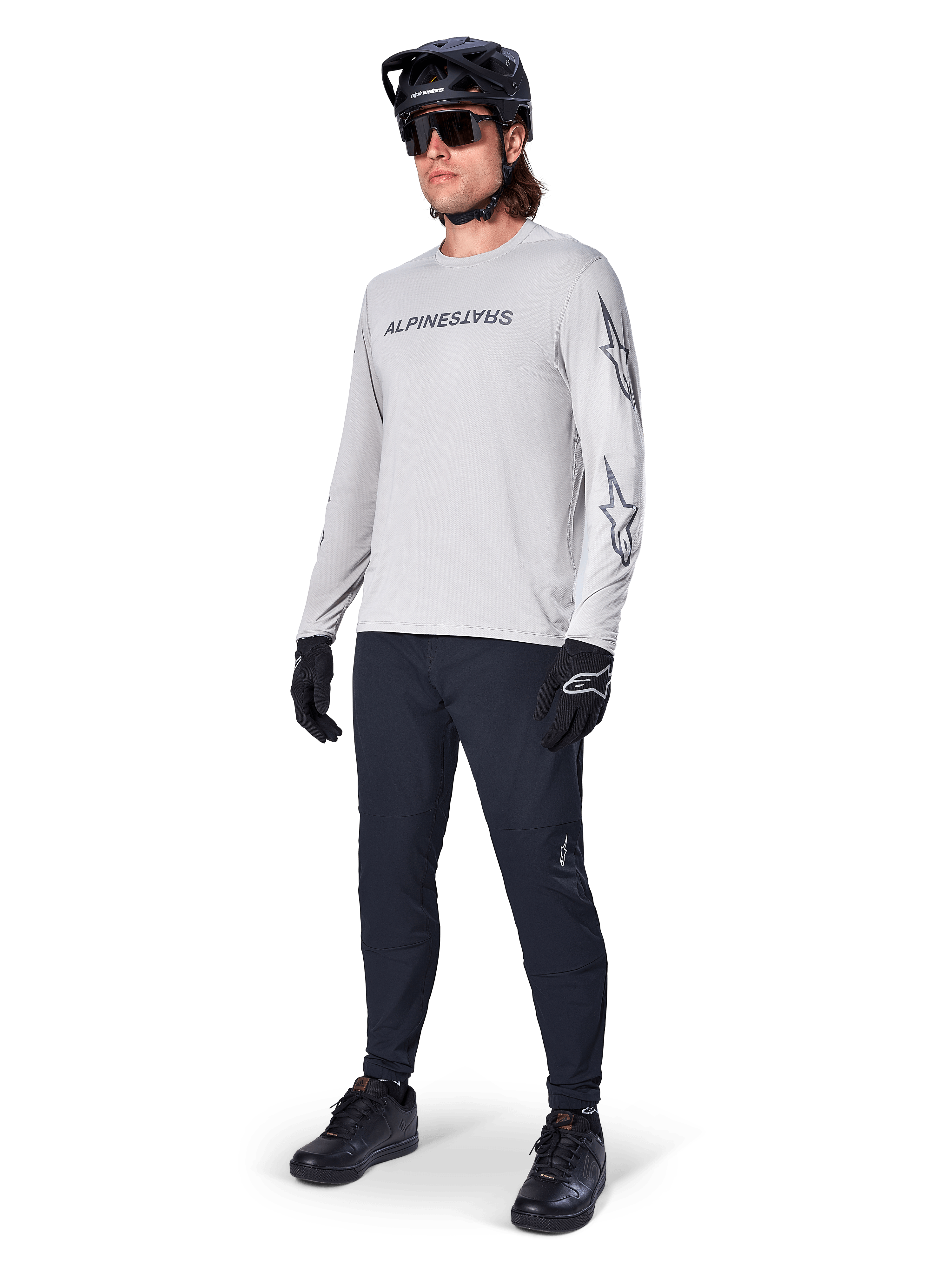Jersey de manga larga Alpinestars A-Dura Switch, gris claro, usado por un ciclista con pantalones negros de montaña y casco, con logotipo de la marca en el pecho y gráficos de estrellas en las mangas, equipamiento de alto rendimiento para ciclismo todoterreno